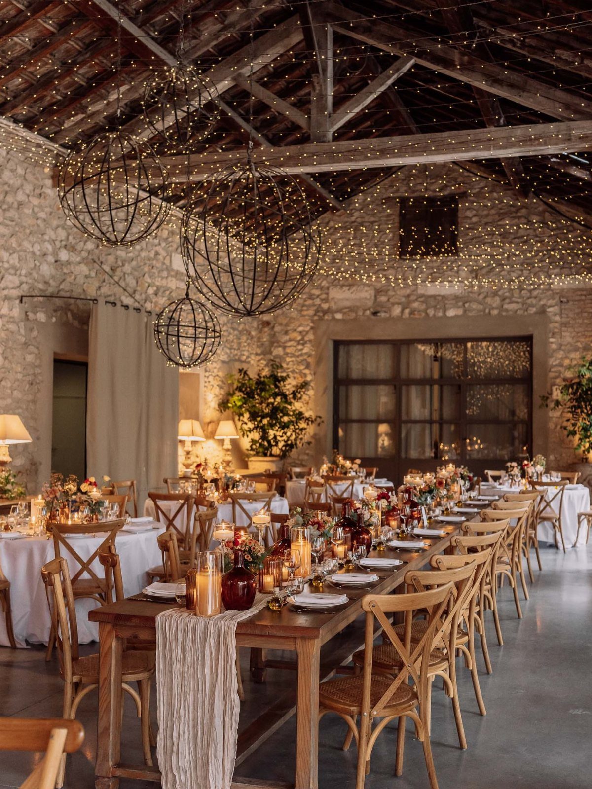 Long wooden banquet tables set with candles and flowers inside a rustic stone building.