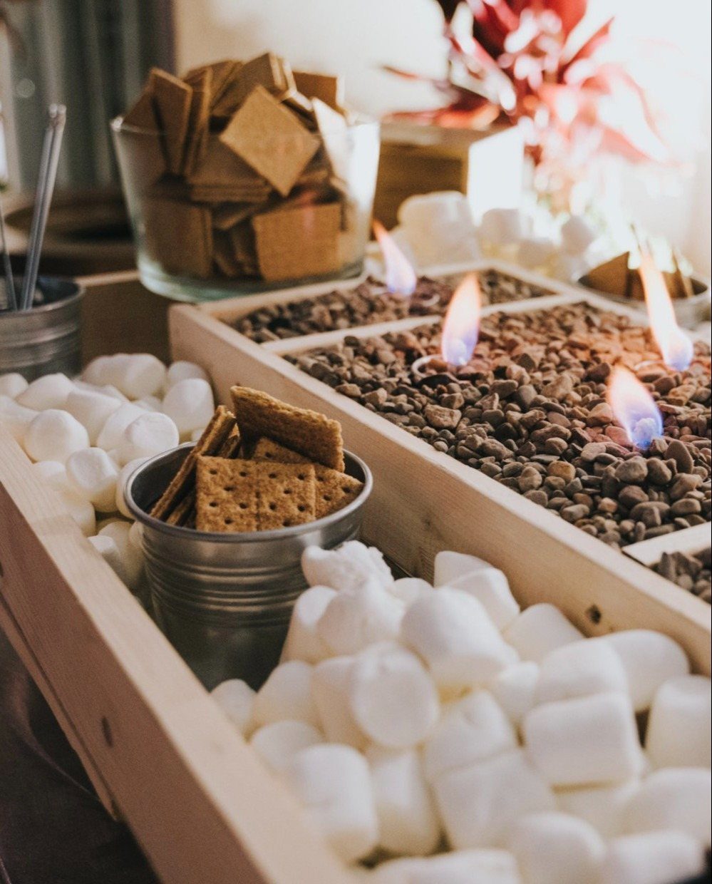 A s'mores station with marshmallows, graham crackers, and small flames in a wooden tray.