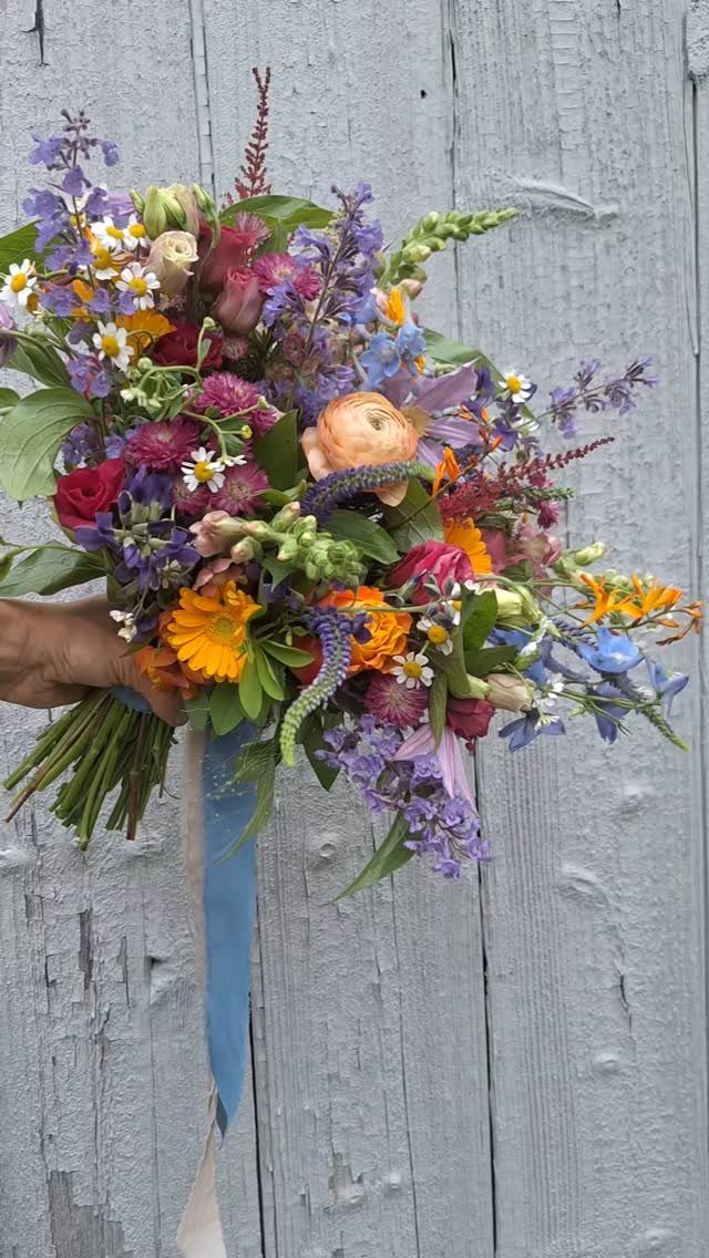 A colorful wildflower bouquet with purple and orange blooms held against a rustic wooden wall.