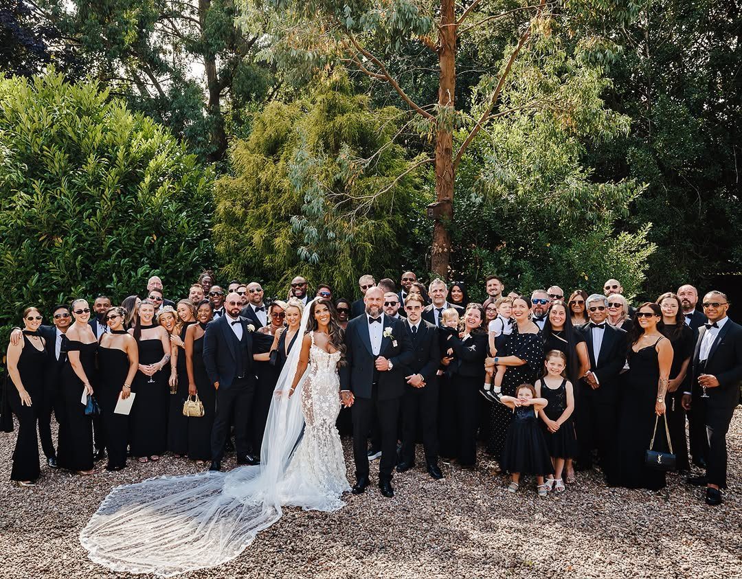 12 Wedding Groom Photos to Capture: From Getting Ready to Golden Hour 5 A large wedding party in formal black attire poses together outdoors in front of lush green trees.