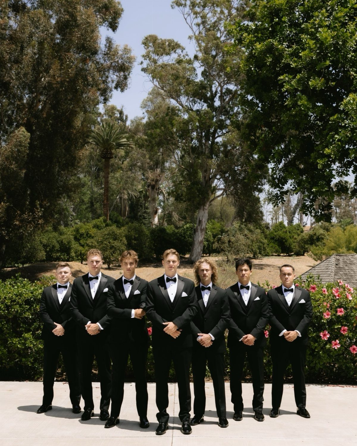 12 Wedding Groom Photos to Capture: From Getting Ready to Golden Hour 12 Seven men wearing black tuxedos standing in a row against a backdrop of green trees.