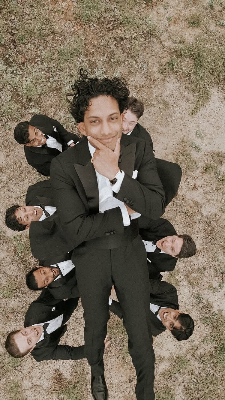12 Wedding Groom Photos to Capture: From Getting Ready to Golden Hour 11 A man in a black tuxedo posing in a circle with his groomsmen on grass.