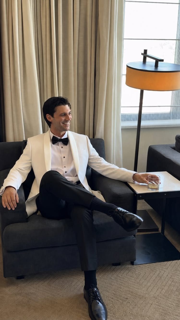 12 Wedding Groom Photos to Capture: From Getting Ready to Golden Hour 7 A smiling man in a white tuxedo jacket sitting in a dark armchair holding a drink.