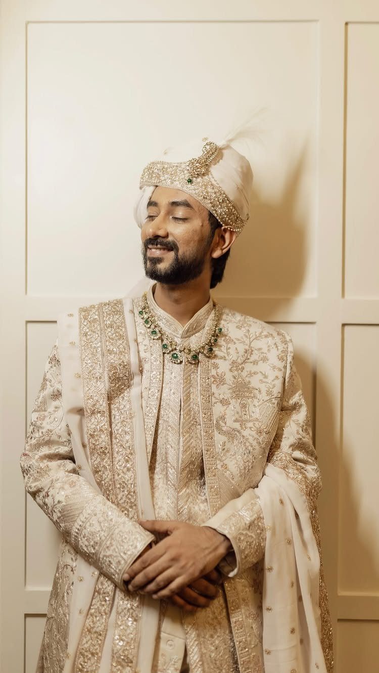 12 Wedding Groom Photos to Capture: From Getting Ready to Golden Hour 9 A man in traditional cream Indian wedding attire and turban standing against a paneled wall.