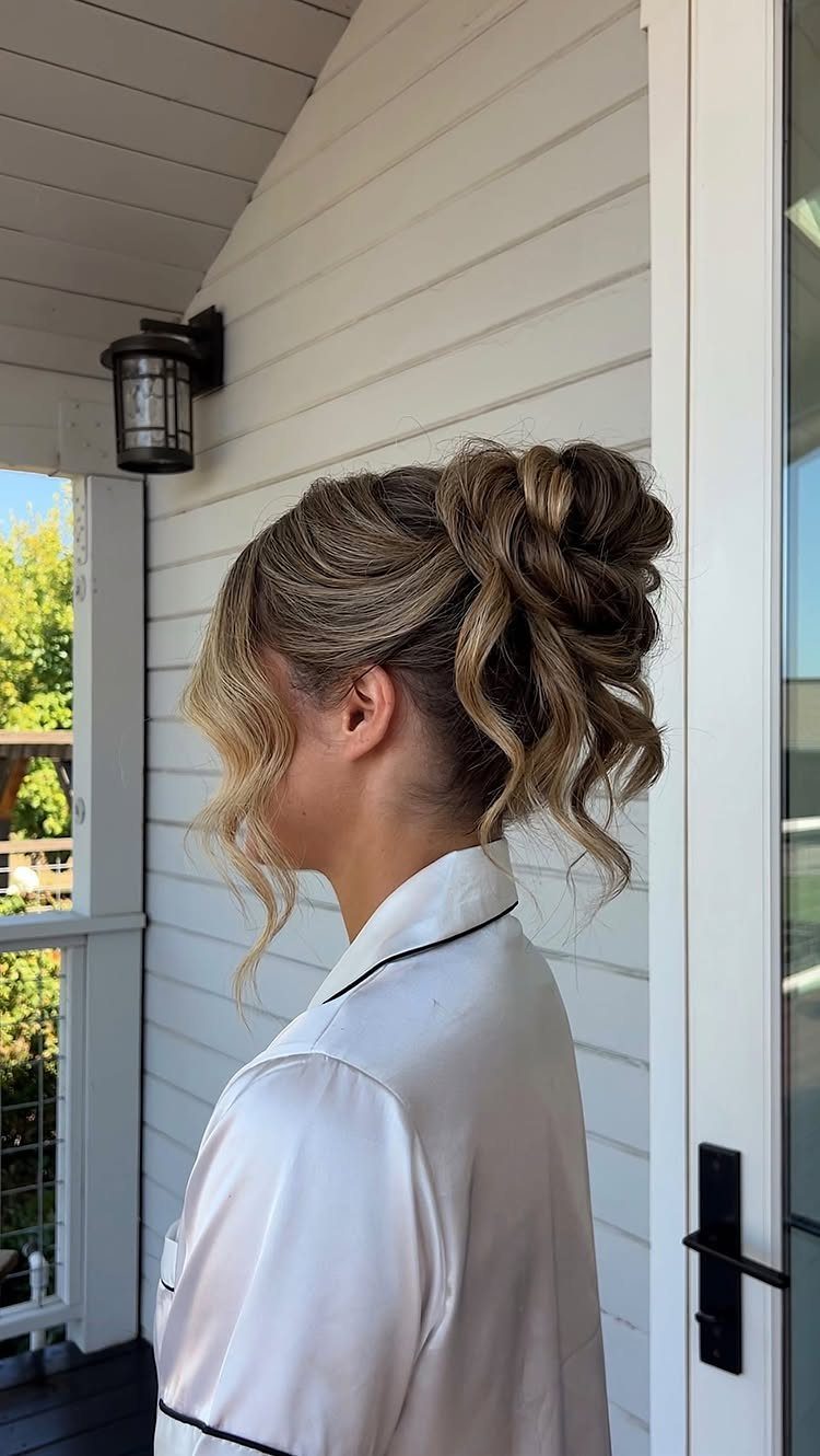 5 Wedding Hair Inspo Ideas for Brides From Loose Curls to Hollywood Waves 5 Woman with blonde highlighted hair in an elegant, wavy bridal updo wearing a white satin robe.
