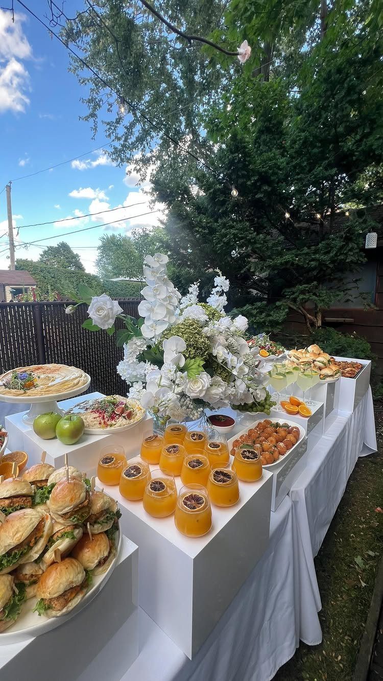 An outdoor buffet table displays sliders, juices, and white flowers under a bright blue sky.