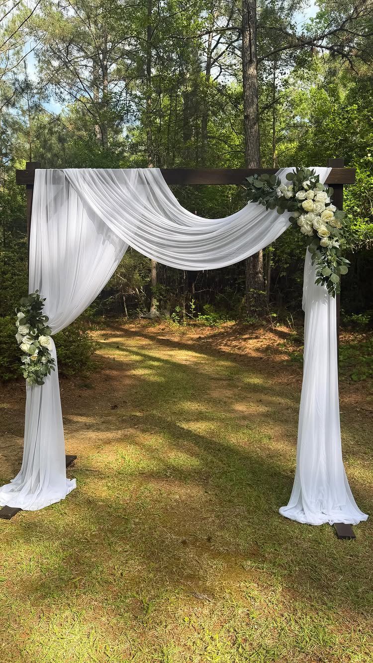 A wooden wedding arch draped in white fabric and adorned with white roses on a lawn.
