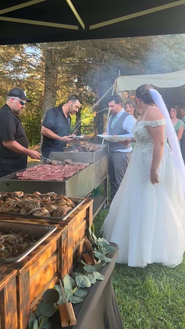 A bride stands near a catering station where chefs prepare grilled meat outdoors.
