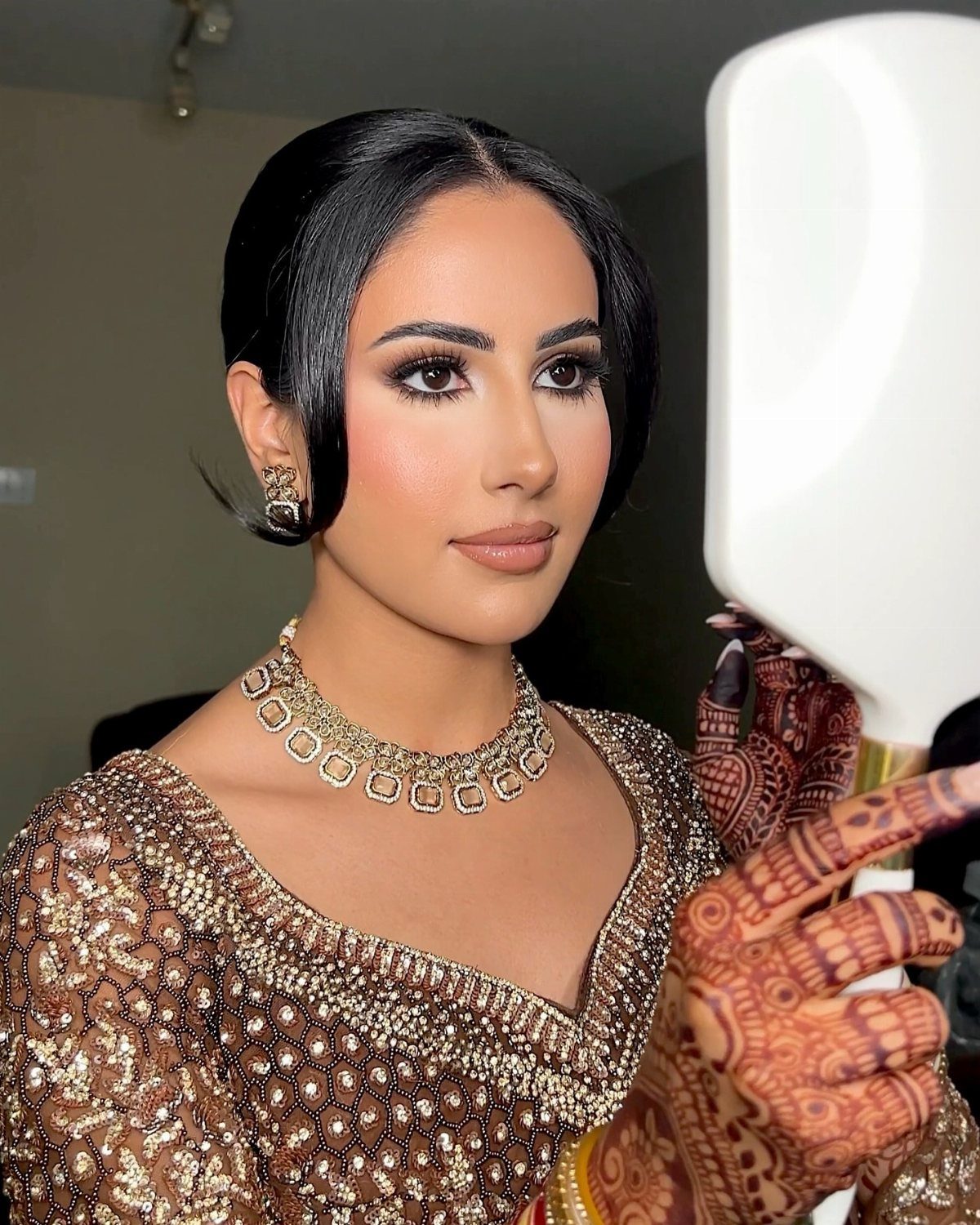 A woman with bronzed makeup and gold jewelry looking into a white vanity mirror.