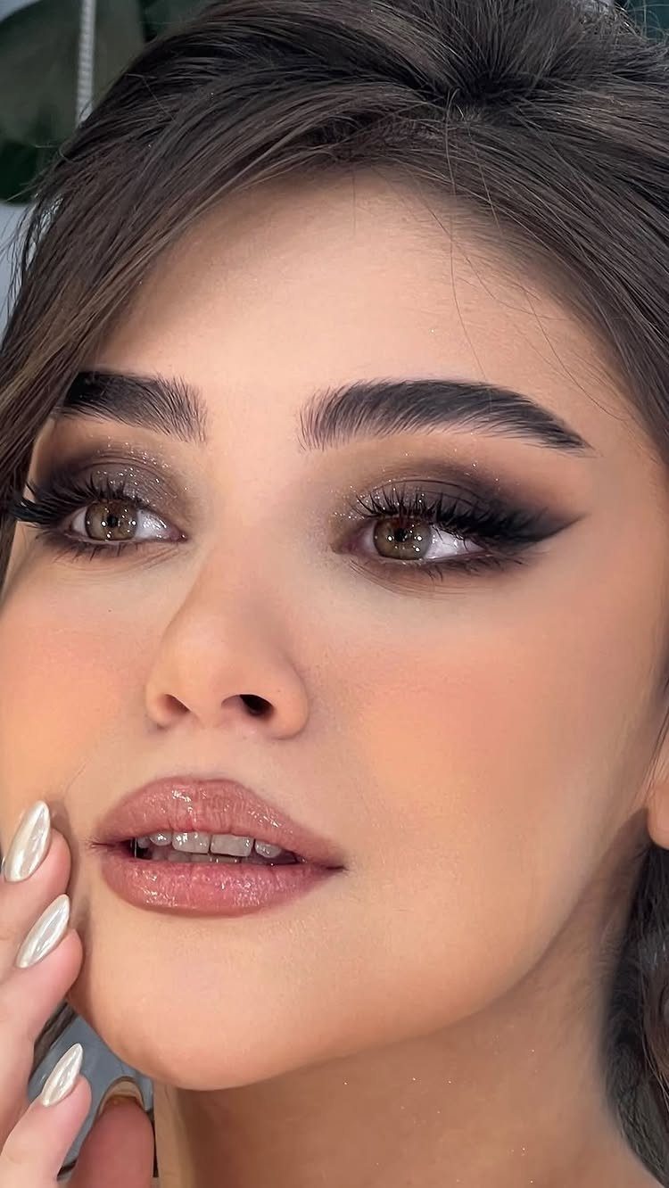 A woman with dramatic winged eyeliner, shimmering eyeshadow, and glossy pink lips looking slightly away.