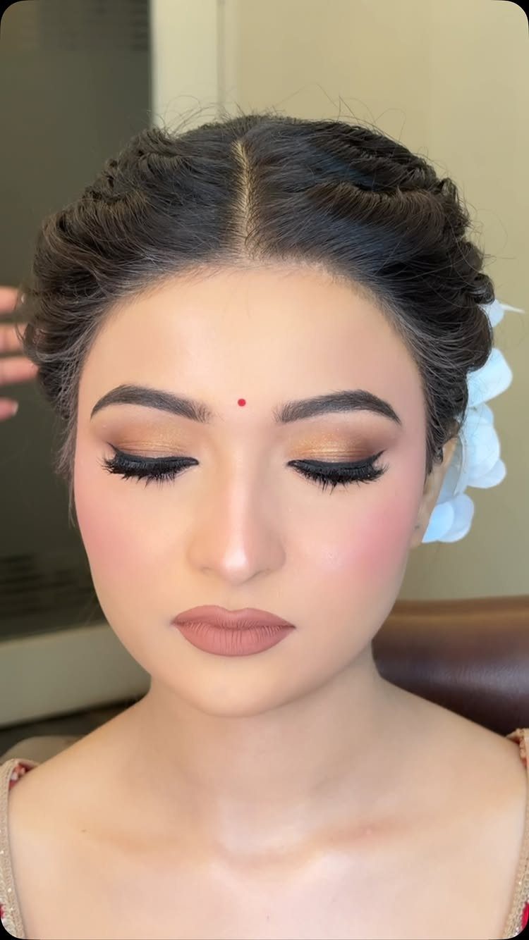 A woman with gold eyeshadow, rosy blush, and braided hair decorated with white flowers.