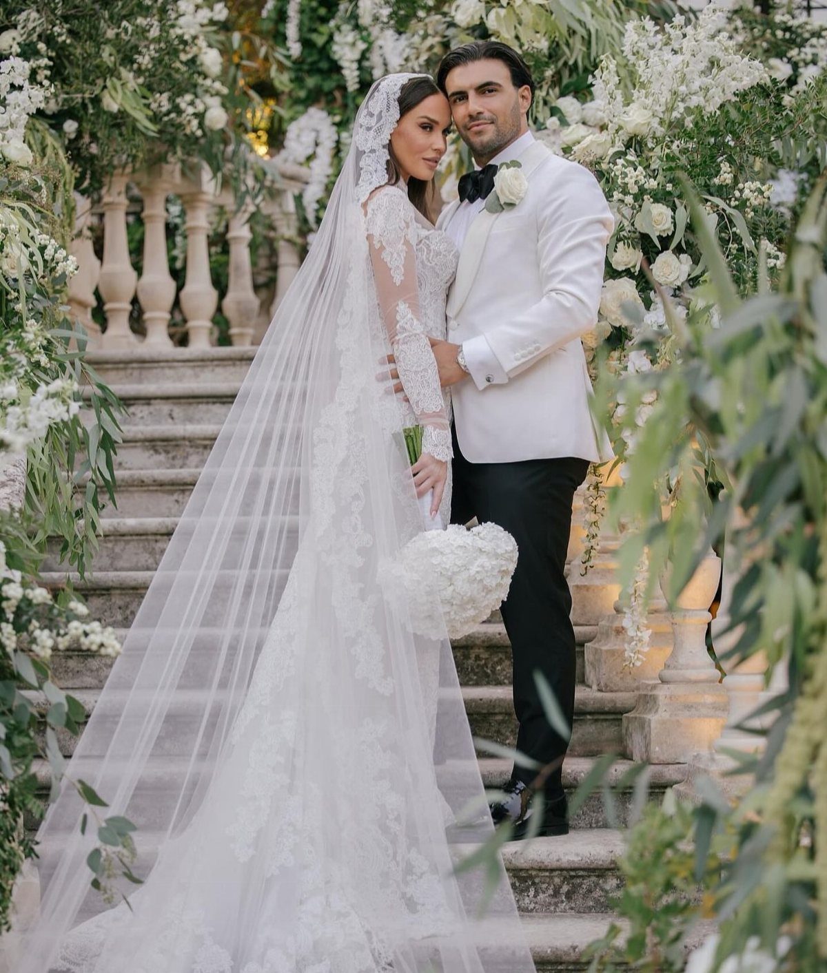 Wedding Outfit for Europe: 6 Ideas for 2026 Ceremonies Abroad 5 A bride in lace and a groom in a white tuxedo pose on stone stairs amidst white flowers.
