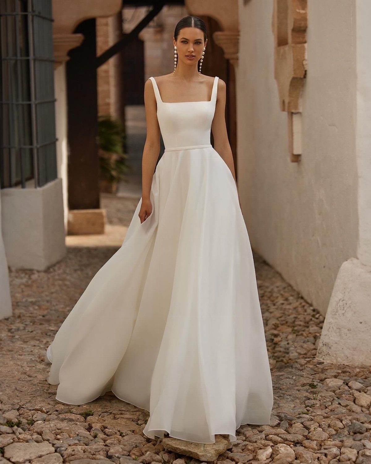 Wedding Outfit for Europe: 6 Ideas for 2026 Ceremonies Abroad 1 A woman in a white satin wedding gown standing on a cobblestone path outdoors.
