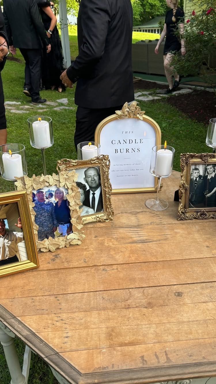 Lit white pillar candles in glass holders and framed portraits on a wooden table outdoors.