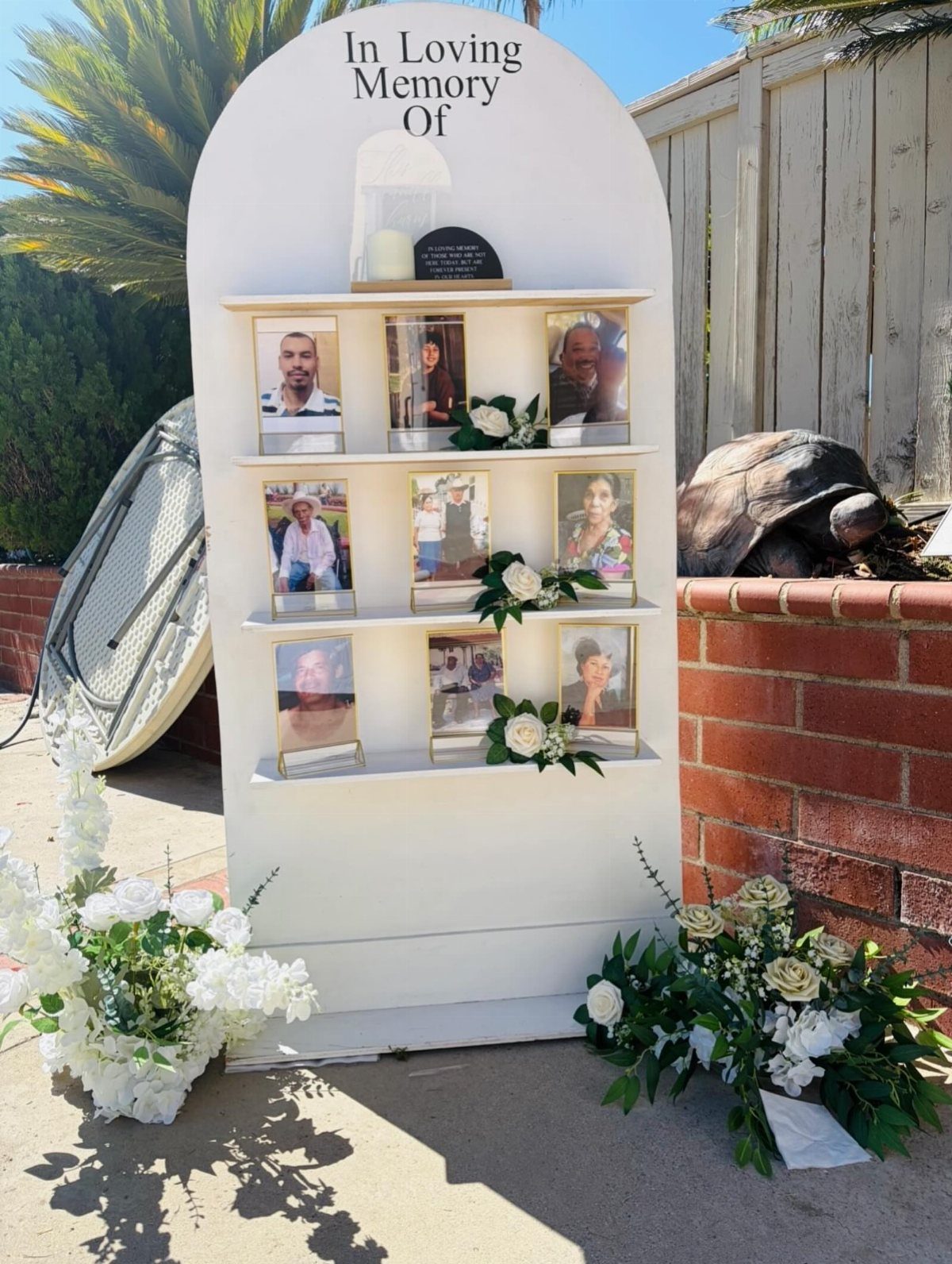 A white arched memorial stand displaying framed photographs and white flowers outdoors.