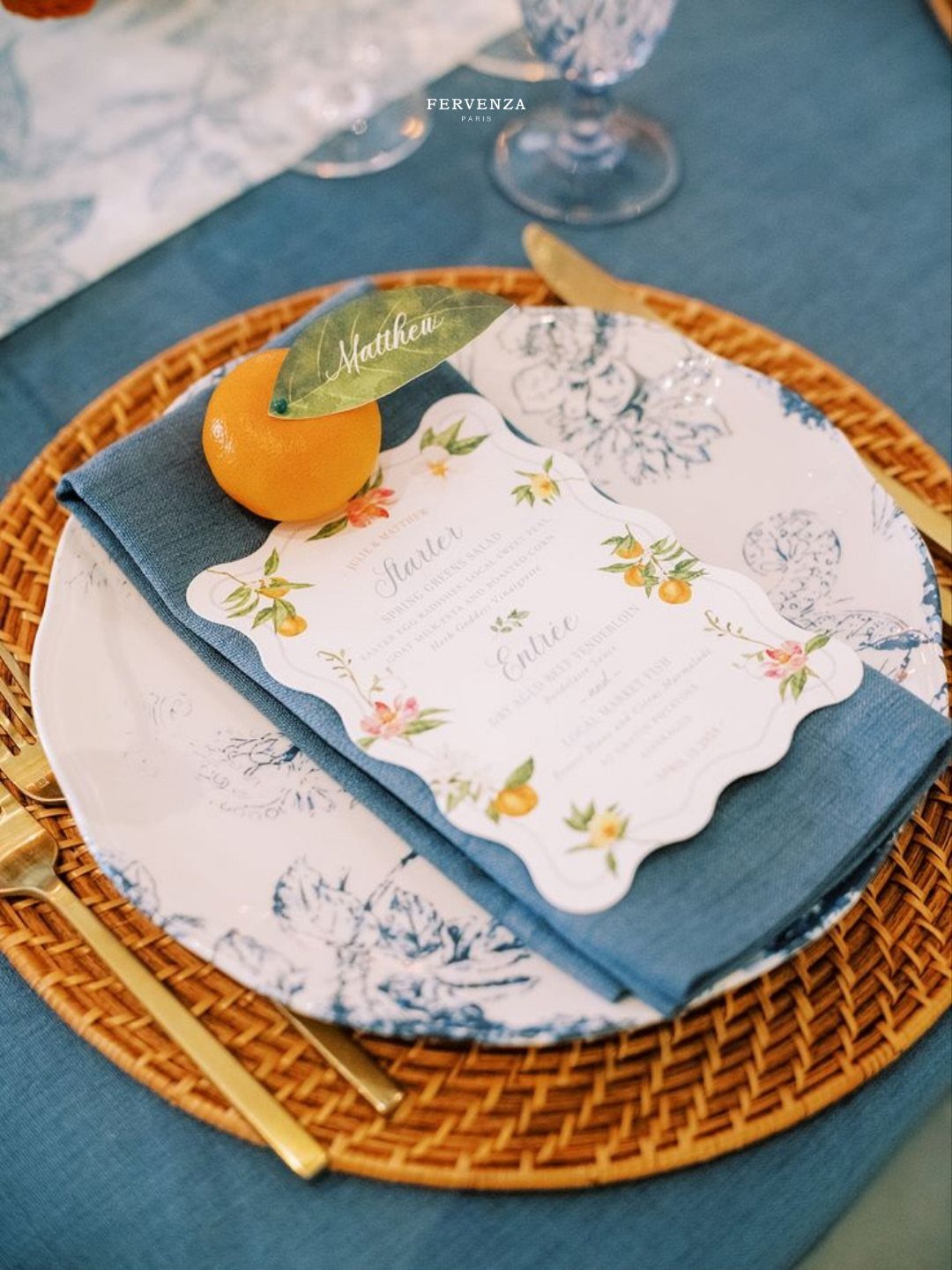 A wedding table setting with blue linens, a woven charger, and a citrus-themed menu card.