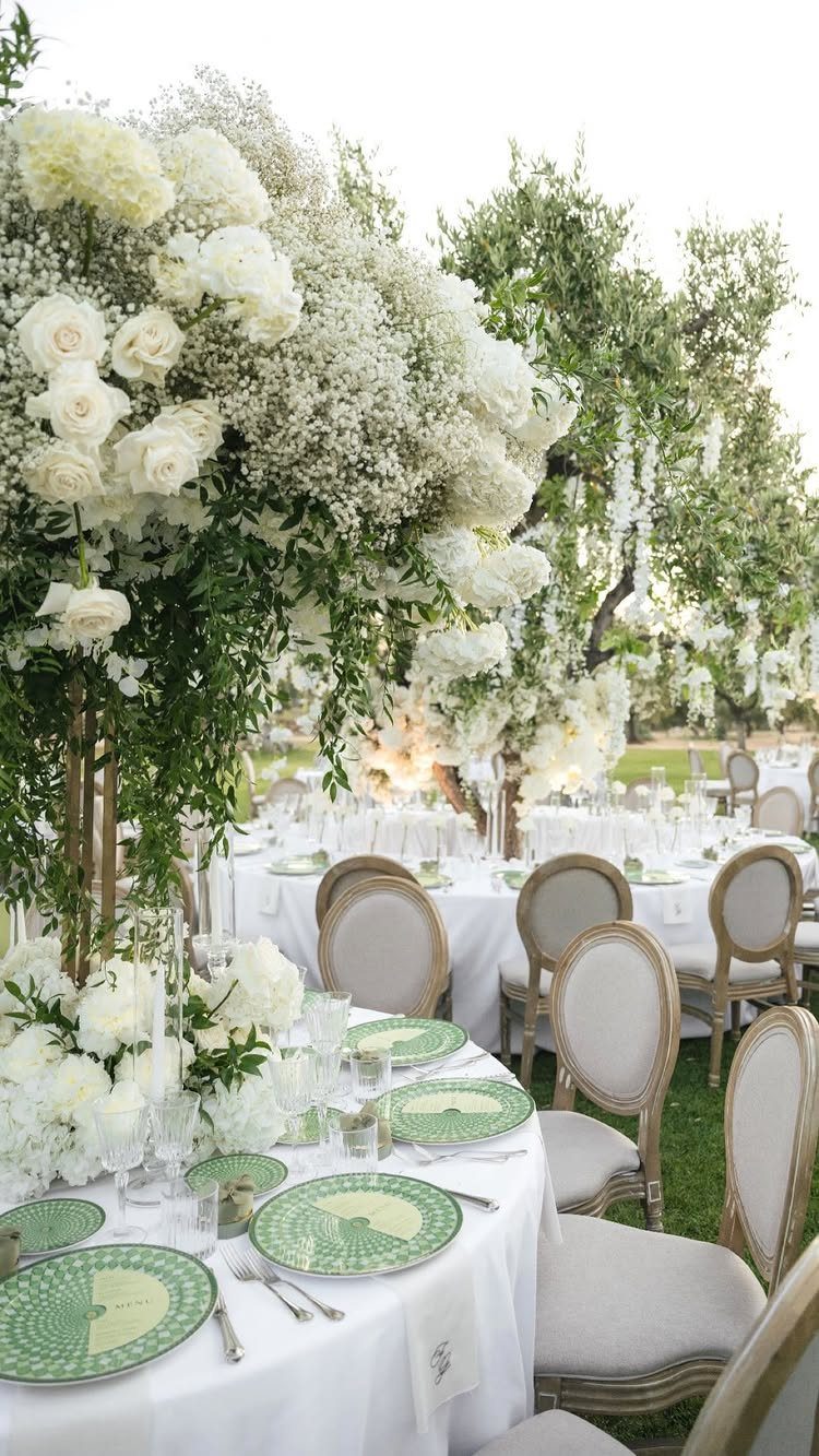 White floral centerpieces and green patterned plates on a wedding reception table outdoors.