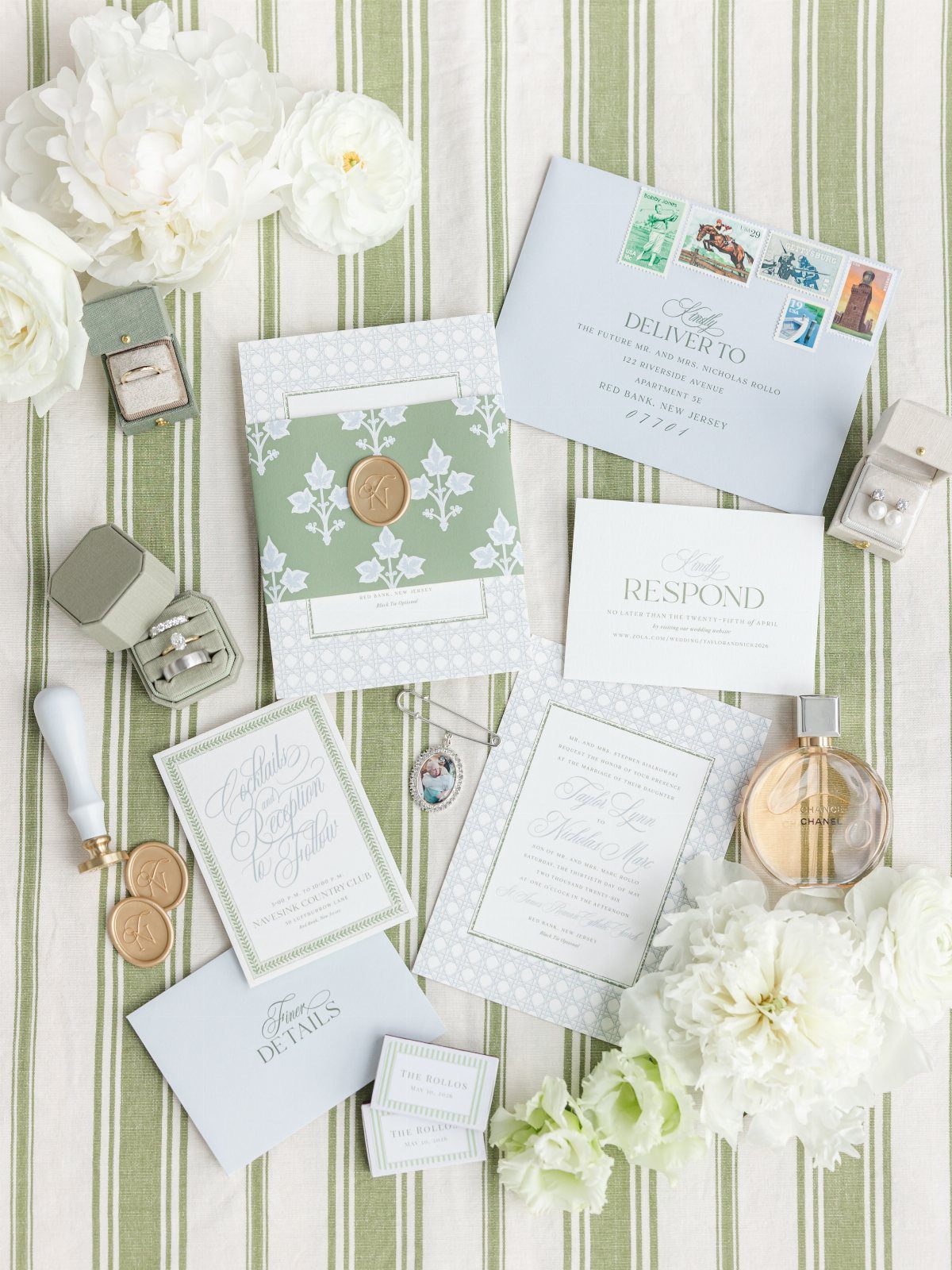 Wedding stationery set with white flowers and jewelry on a green striped surface.