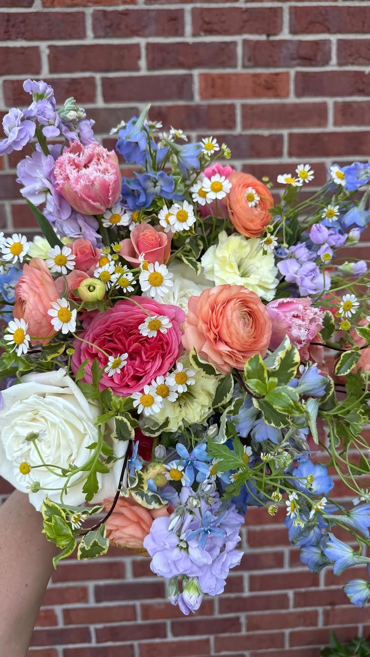A colorful bouquet of pink, peach, and blue flowers with white daisies held against a brick wall.
