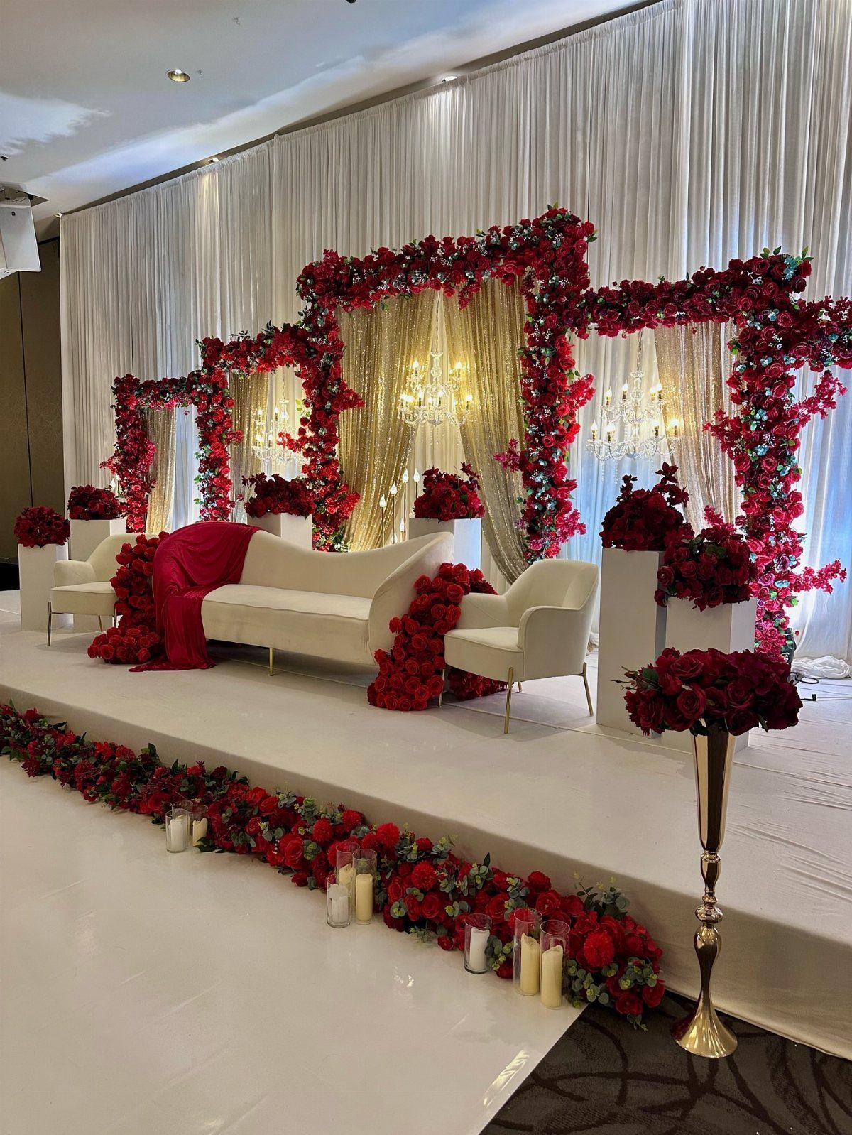 Wedding stage featuring white furniture, gold backdrops, and lush red rose arrangements on a white platform.