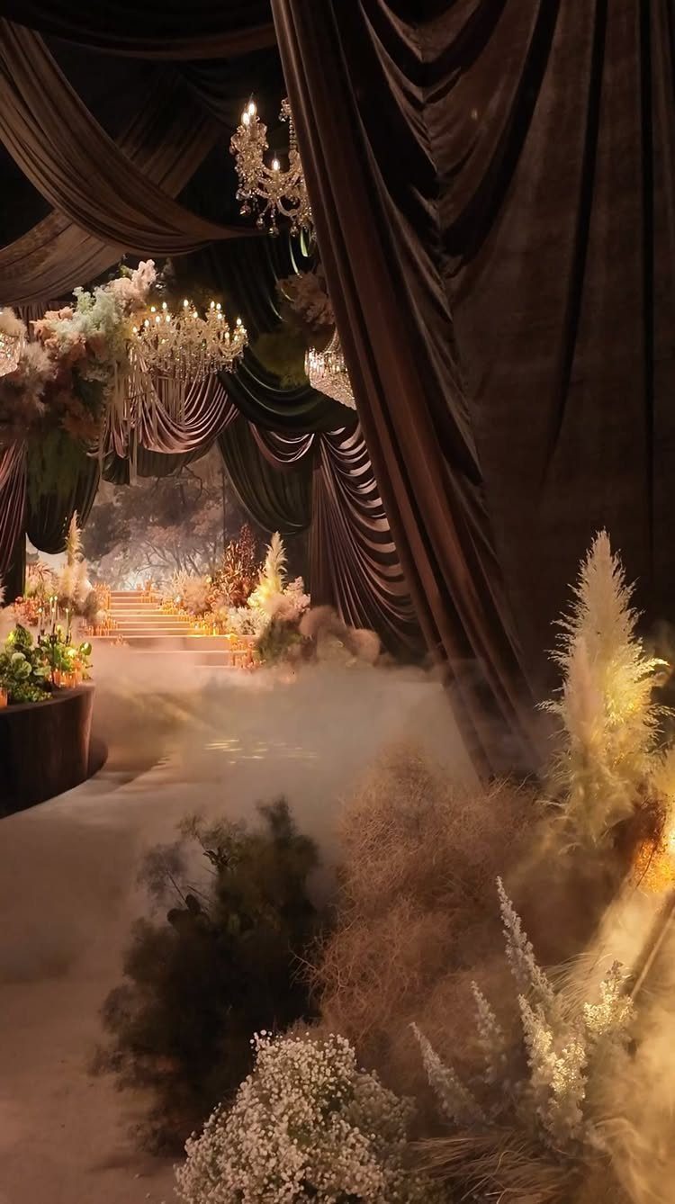 Dark draped fabric and crystal chandeliers illuminate a misty path lined with dried flowers and pampas grass.