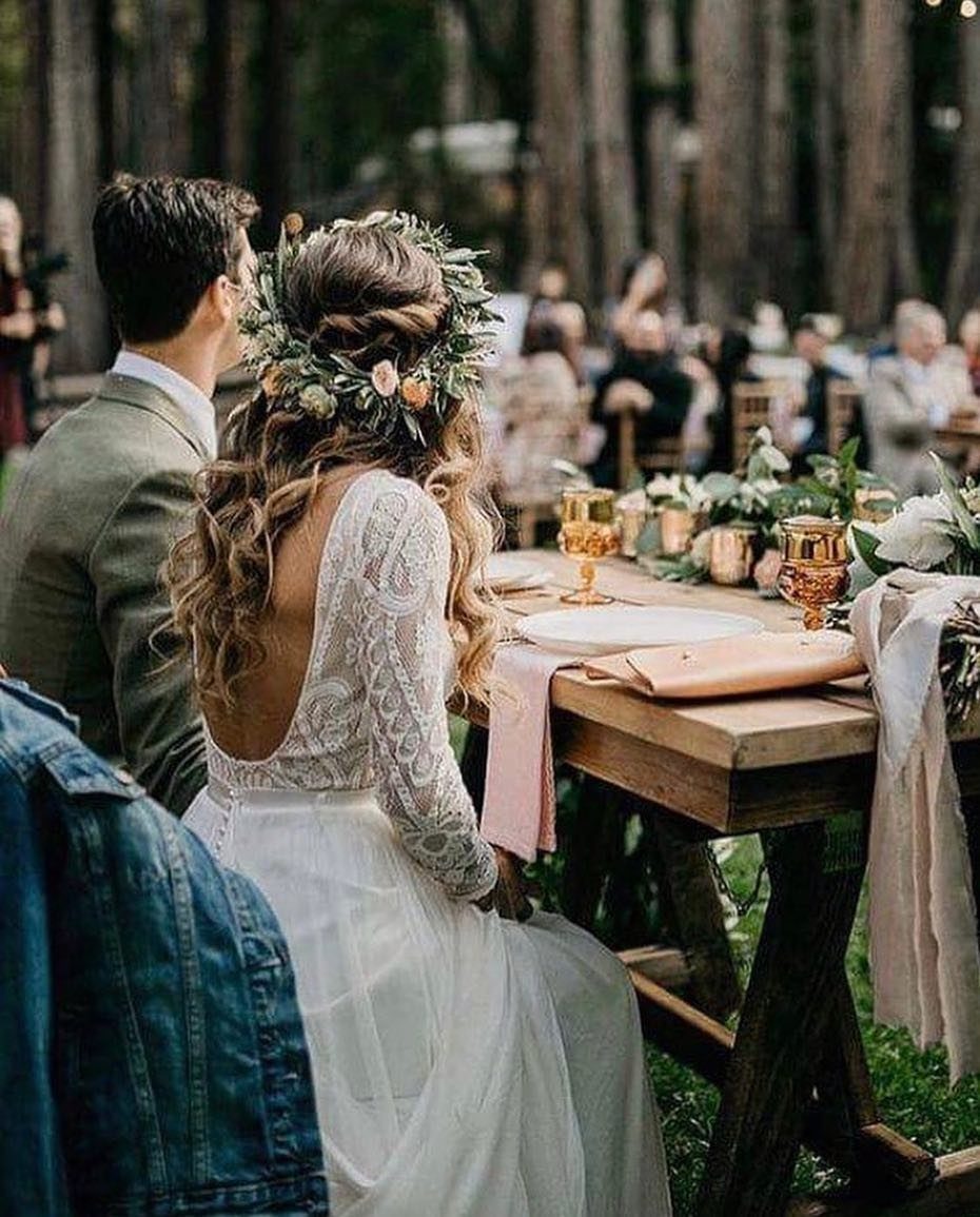 A bride wearing a flower crown and lace dress sits at an outdoor wedding table.