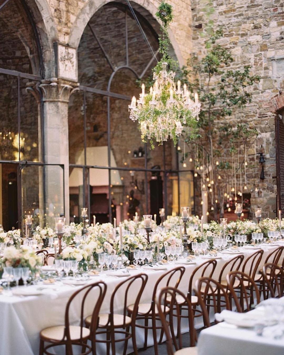 A long wedding banquet table decorated with white flowers and candles inside a stone hall.