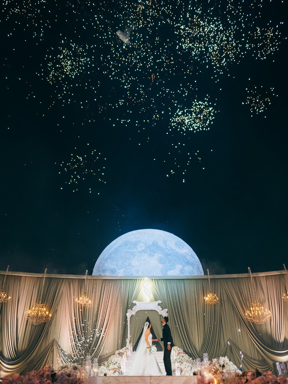 A bride and groom stand before a moon-themed wedding altar under a firework-filled night sky.