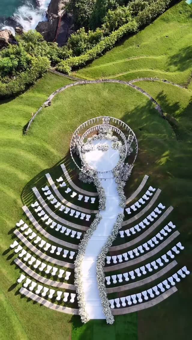 Aerial view of a white wedding ceremony setup on a green lawn by the ocean.