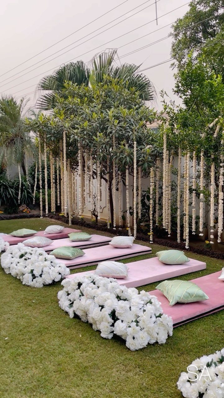 Pink floor cushions with light pillows arranged on grass near white flowers and hanging garlands.