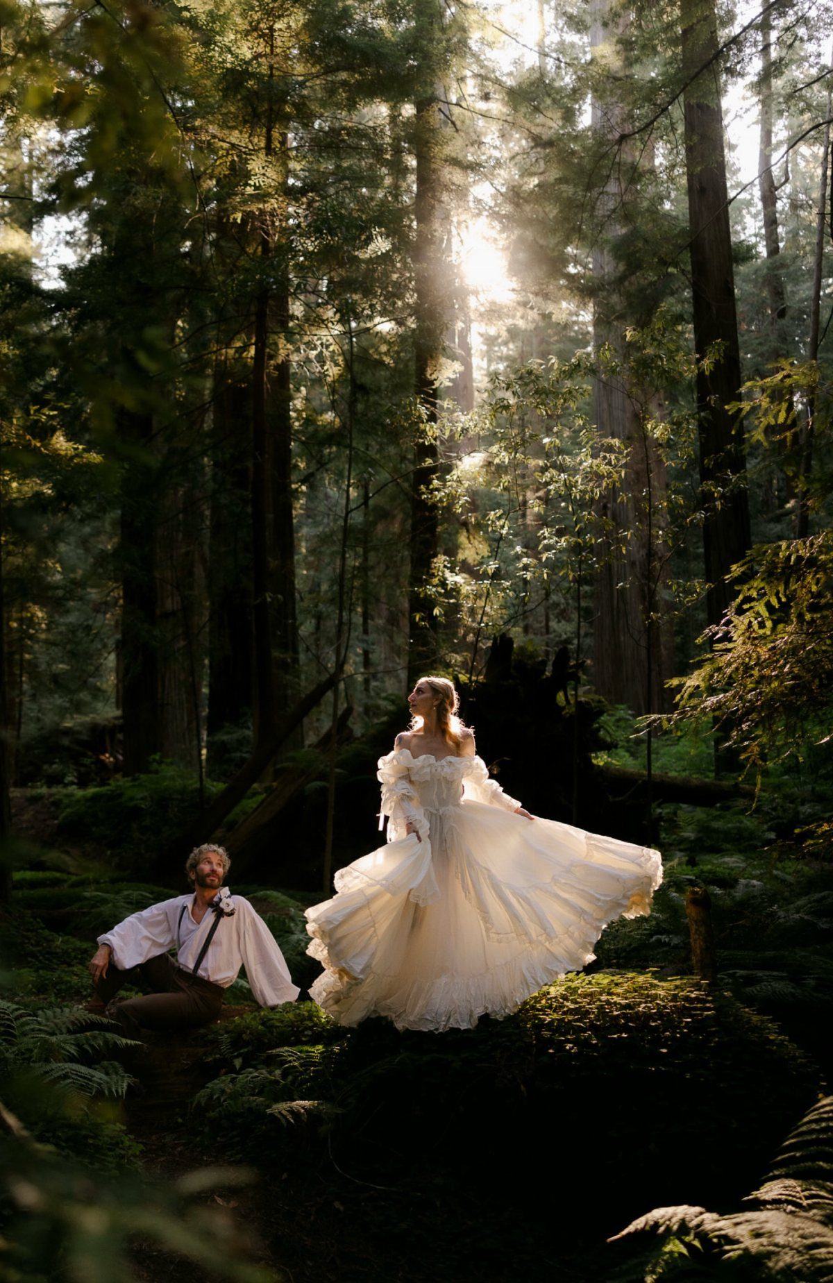 A bride in a flowing white gown and a groom pose in a sunlit, misty forest.