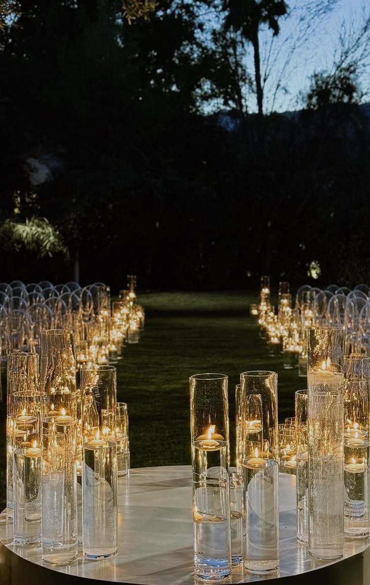Tall glass cylinder vases with floating candles lining a dark outdoor grass path at twilight.