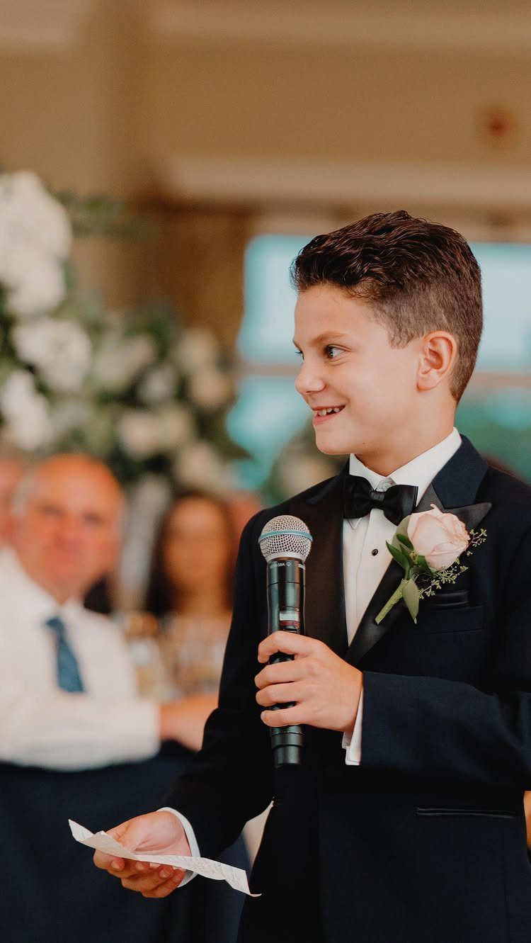A young boy in a tuxedo holding a microphone and speech notes at a wedding.