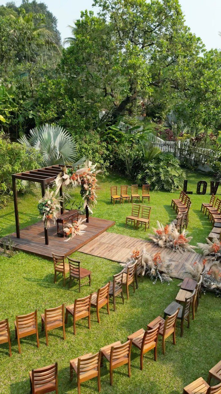 Outdoor wedding ceremony setup with wooden chairs, a pergola, and lush green trees.