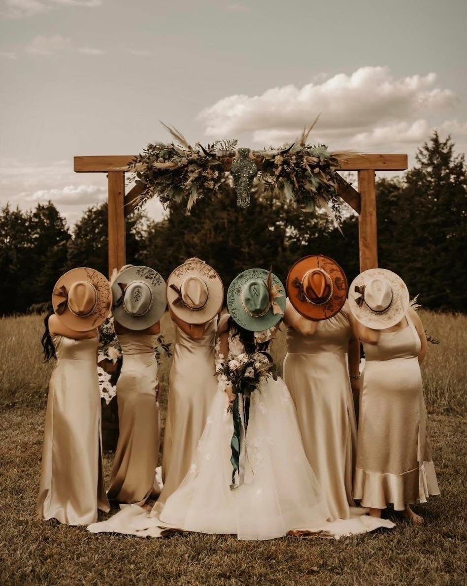Bridesmaids and a bride holding hats in front of a wooden floral wedding arch.