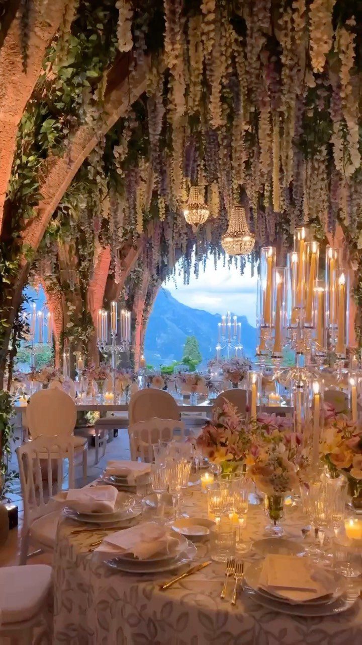 Elegant wedding reception tables set under a lush canopy of hanging wisteria flowers overlooking mountains.