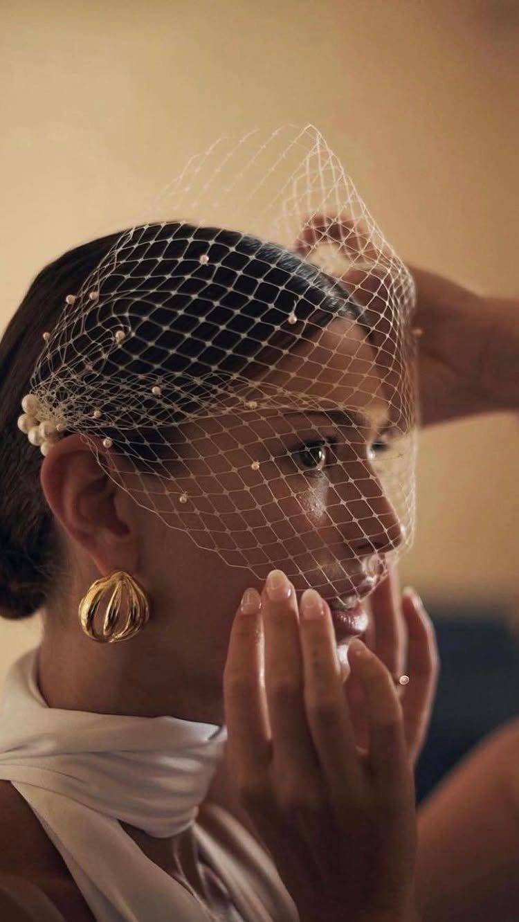 A woman wearing a pearl-encrusted white birdcage veil and gold earrings.
