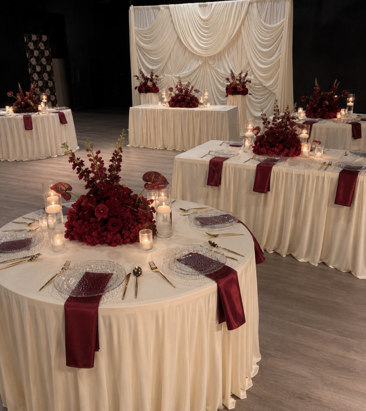 Banquet tables decorated with cream pleated tablecloths, red floral centerpieces, and burgundy napkins in a ballroom.