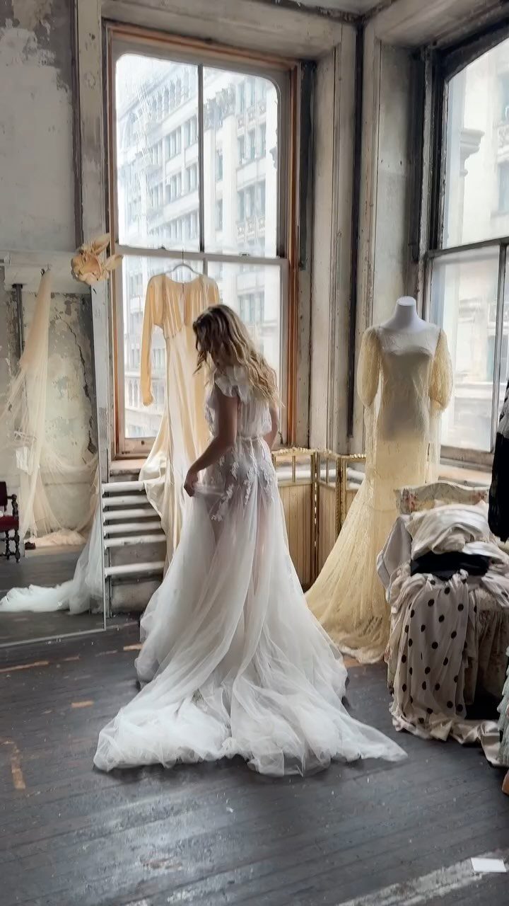 A woman wearing a white lace and tulle wedding dress stands in a sunlit, rustic studio.