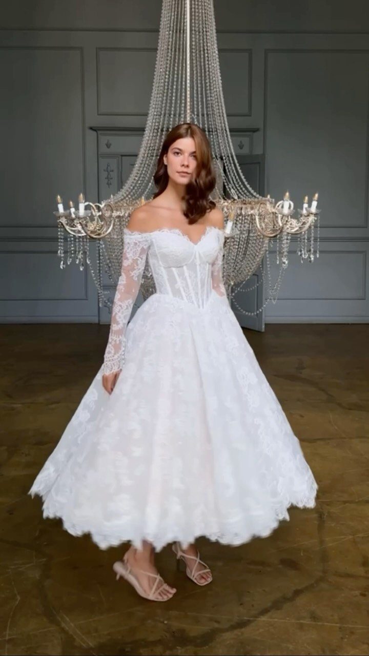 A woman wearing a white lace tea-length wedding dress standing before a large crystal chandelier.