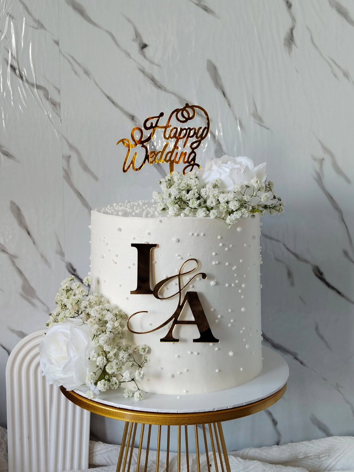 Single-tier white buttercream cake with white roses, baby's breath, and a gold monogram on a gold stand.
