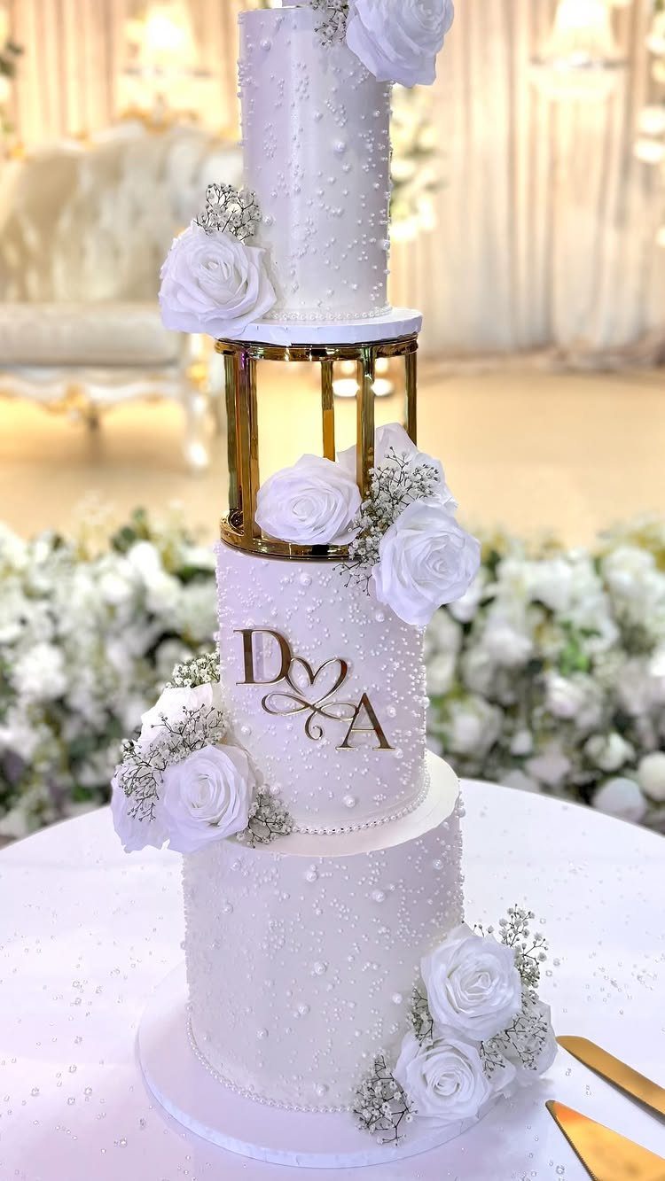 Three-tier white wedding cake decorated with white roses, edible pearls, and gold monogram initials.