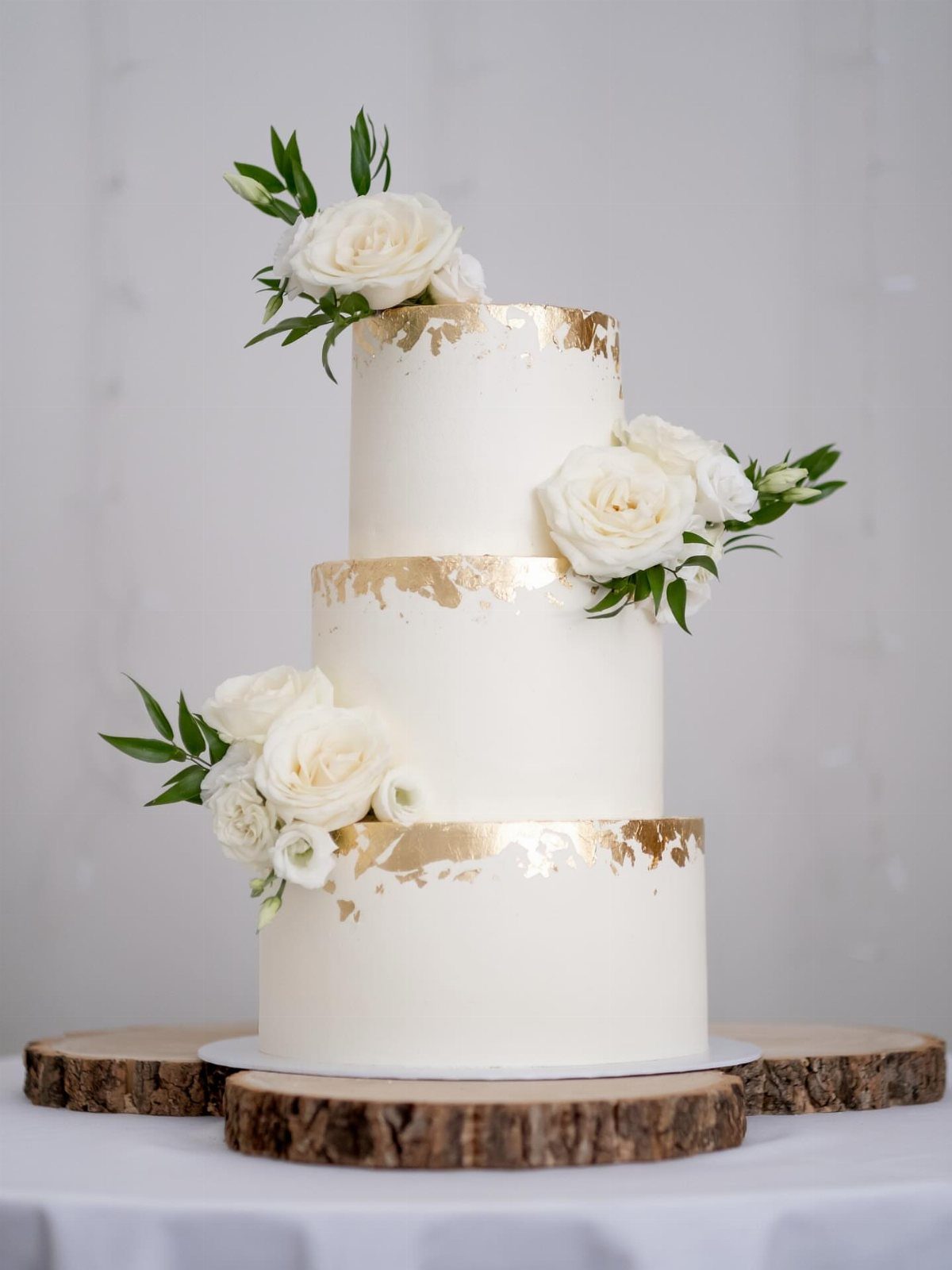 Three-tier white cake with gold leaf accents and white roses on a wooden base.