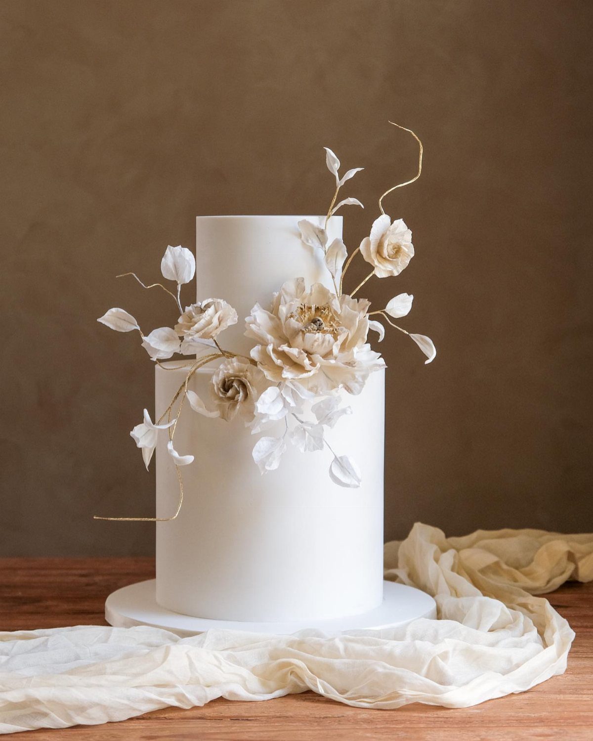 A two-tier white cake decorated with large cream-colored sugar flowers and delicate leaves.