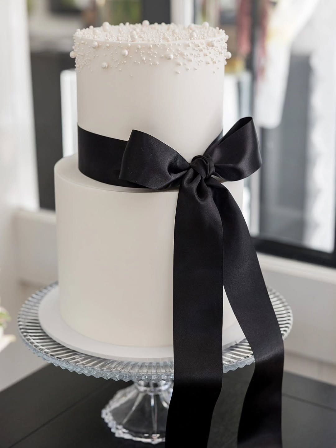 Two-tier white fondant cake decorated with white pearls and a large black satin ribbon bow.