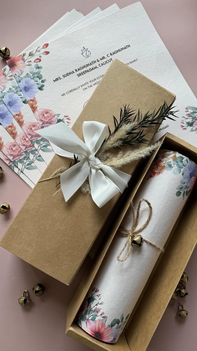 Wedding invitations with watercolor wildflowers, kraft paper packaging, and dried botanical accents on a pink surface.