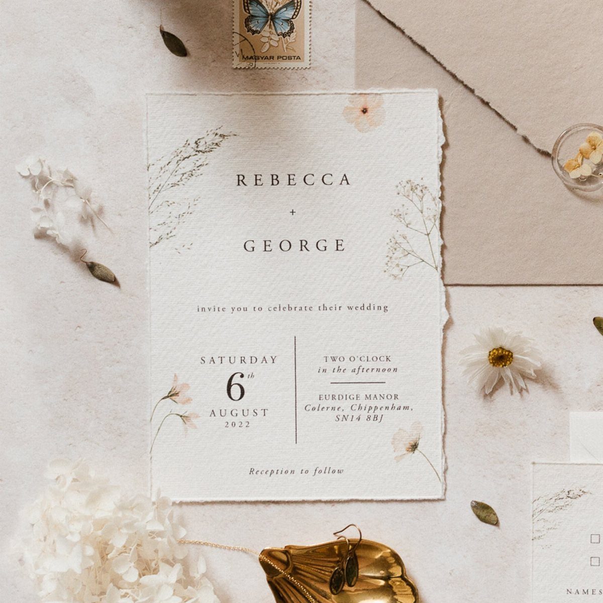 A deckle-edged wedding invitation with wildflower illustrations and elegant text surrounded by dried flowers.