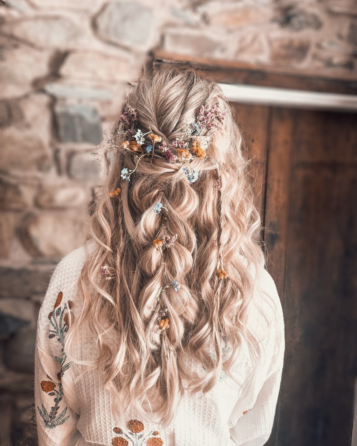 Long wavy blonde hair styled in a braid and decorated with small colorful wildflowers.