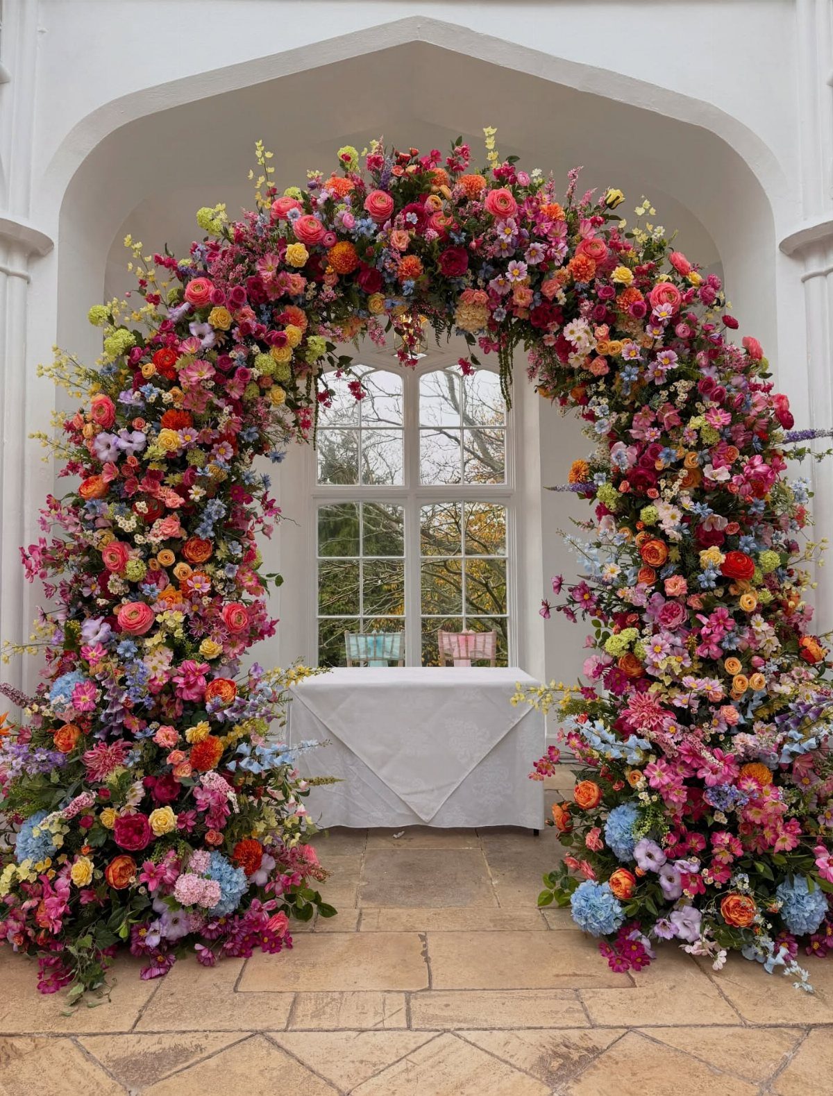 A large, colorful floral archway made of various wildflowers framing a white doorway and table.