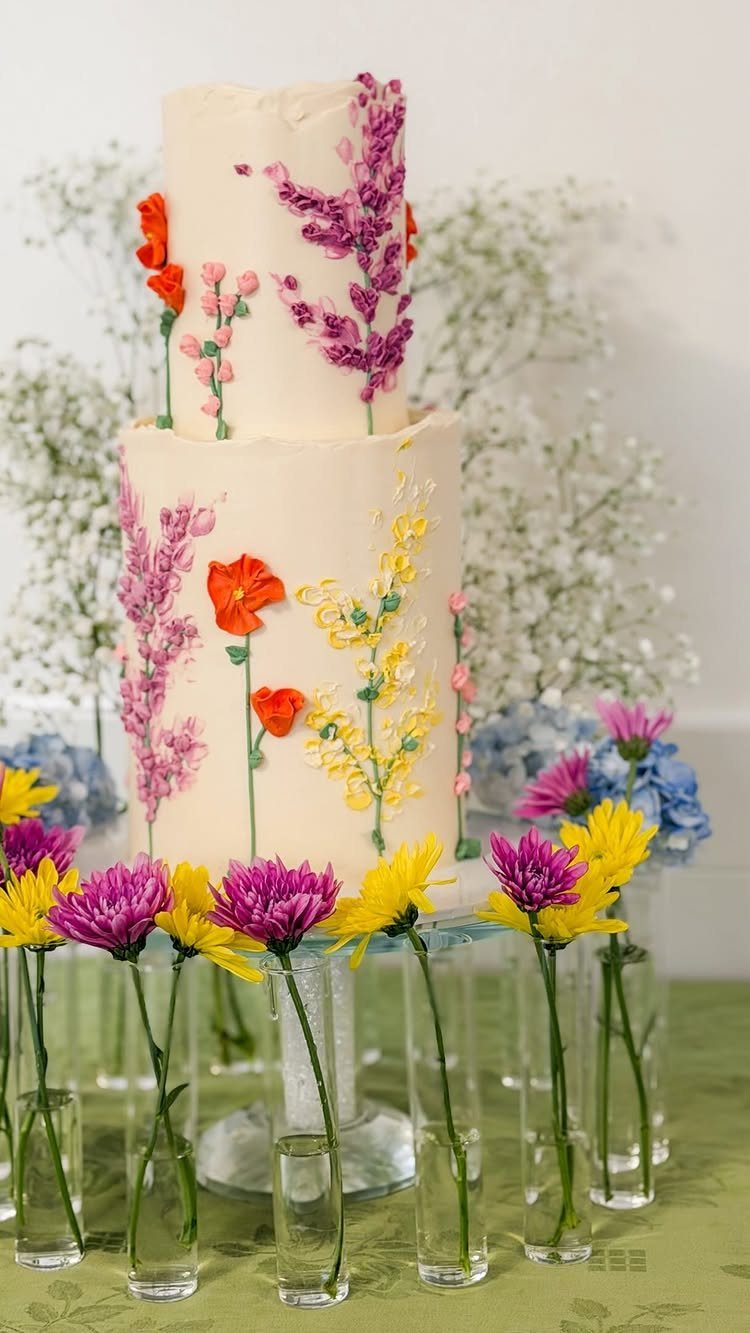 Two-tier white wedding cake decorated with colorful edible wildflowers on a glass stand.